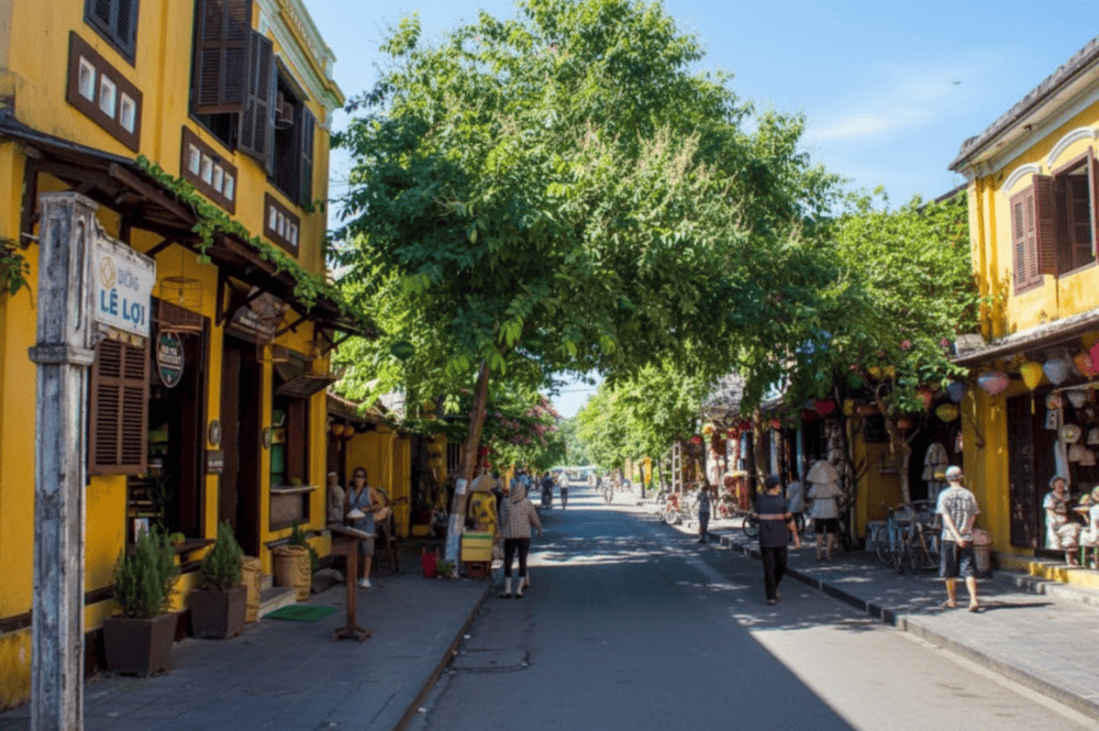 A serene scene on Le Loi Street, Hoi An, with vibrant yellow buildings, greenery, and the lively flow of local life